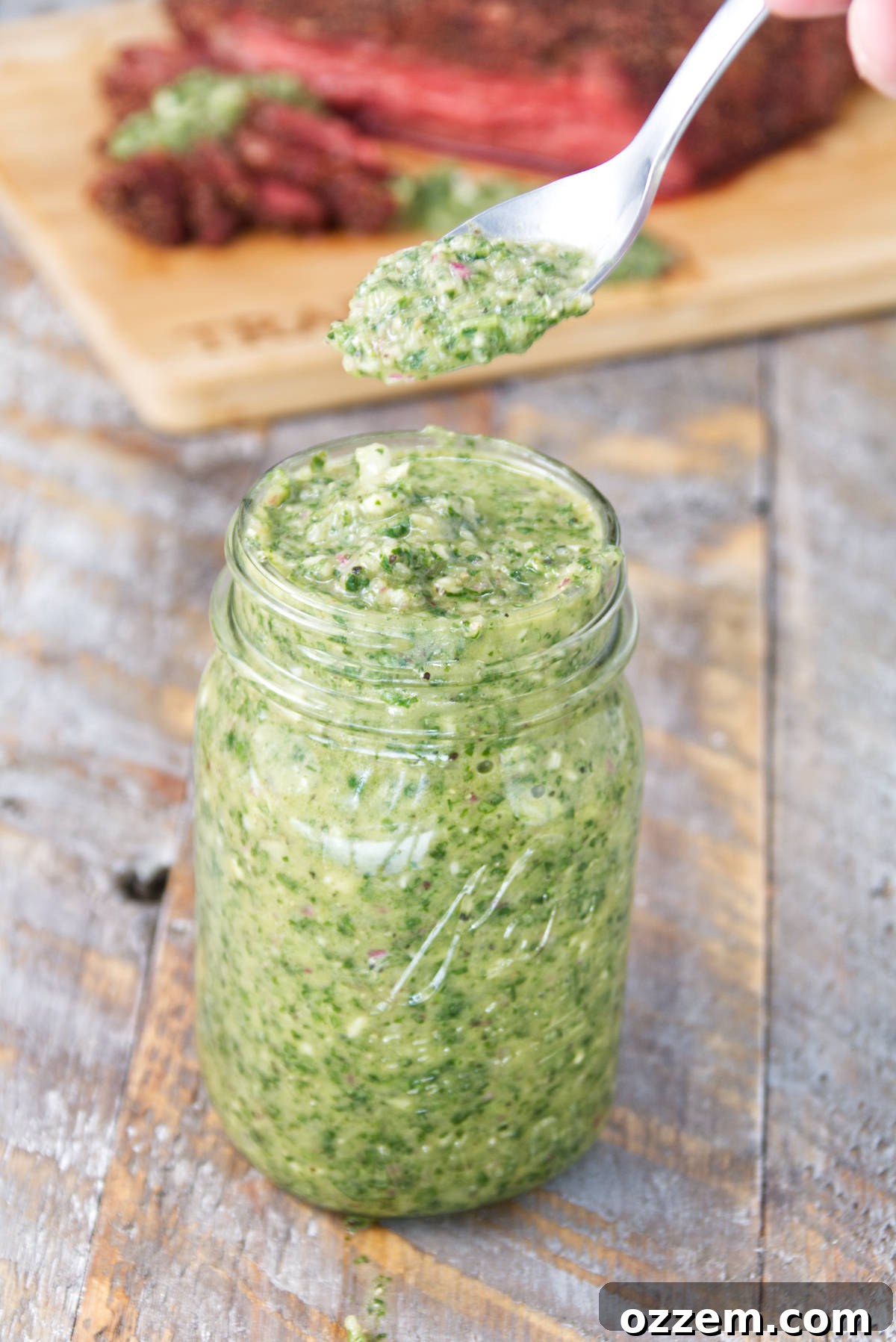 A mason jar brimming with vibrant green mint cilantro chimichurri sauce, set against a backdrop of perfectly sliced smoked tri-tip on a rustic cutting board, showcasing its rich texture and appetizing appeal.