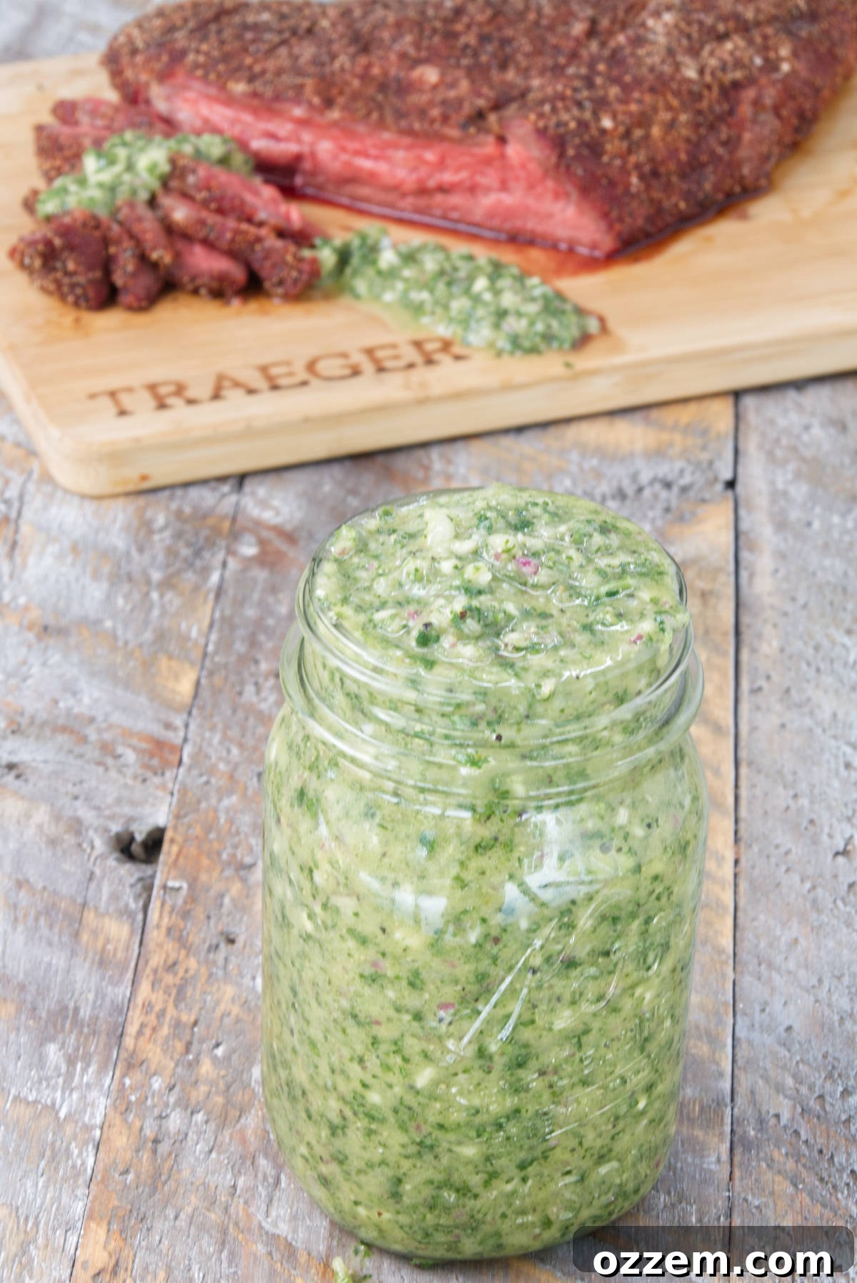 A beautifully presented mason jar filled with vibrant green cilantro chimichurri sauce, with a cutting board displaying sliced smoked tri-tip in the softly blurred background.