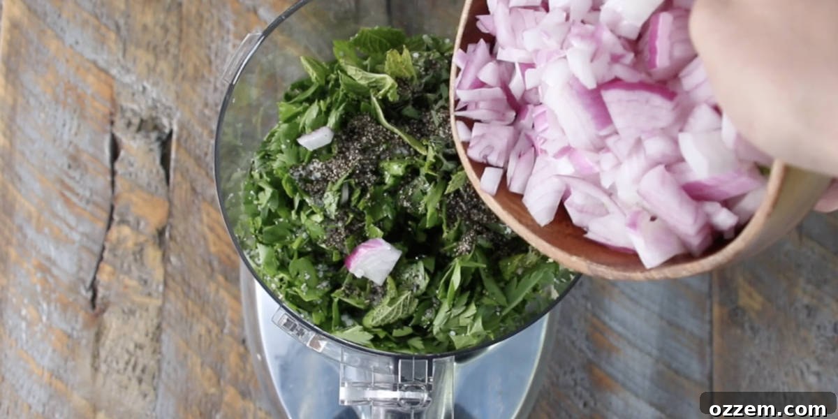 Fresh chimichurri ingredients, including herbs, garlic, and onion, being carefully added to a food processor bowl before blending.