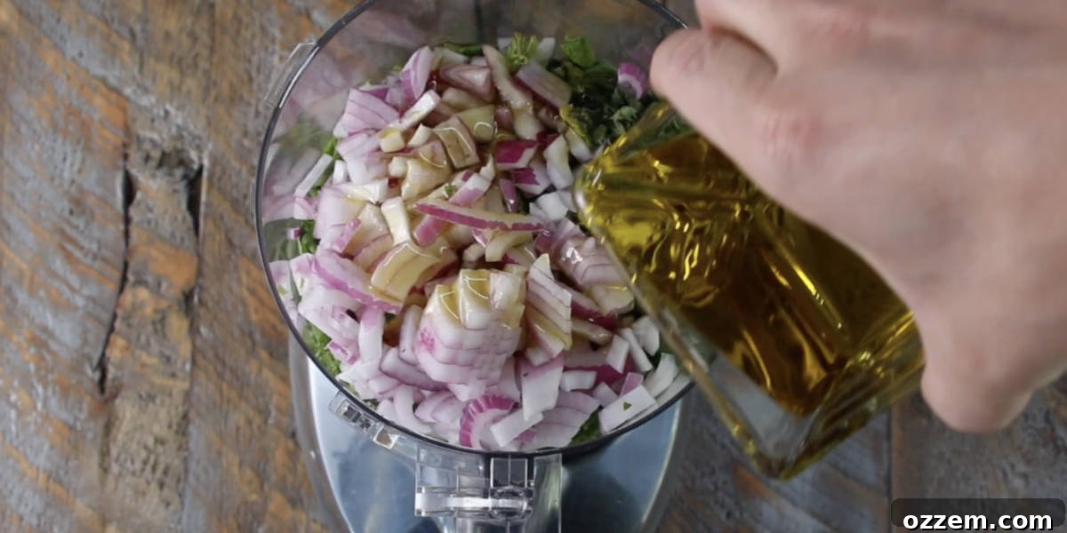 Half of the olive oil being poured into the food processor, mixing with the chimichurri ingredients already inside, before the final blending stage.
