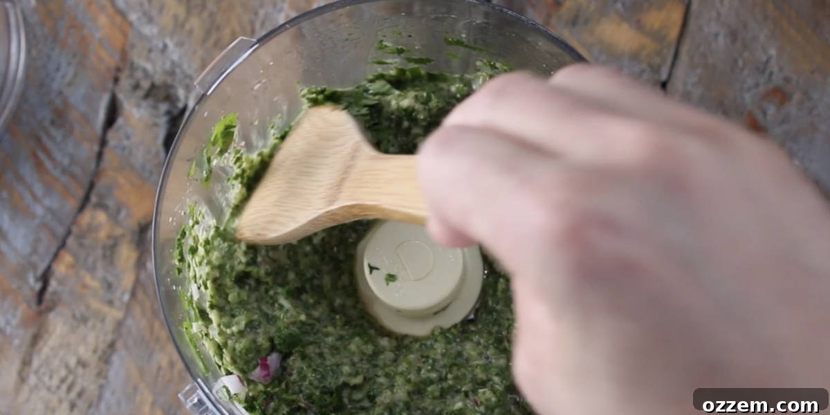 A spatula scraping down the vibrant green chimichurri mixture from the sides of a food processor bowl after initial pulsing, ensuring even blending.