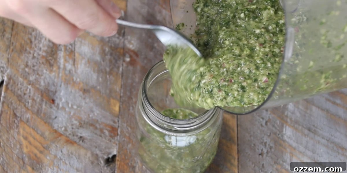 Freshly blended chimichurri sauce being poured from a food processor into a glass mason jar, ready for storage or immediate use.