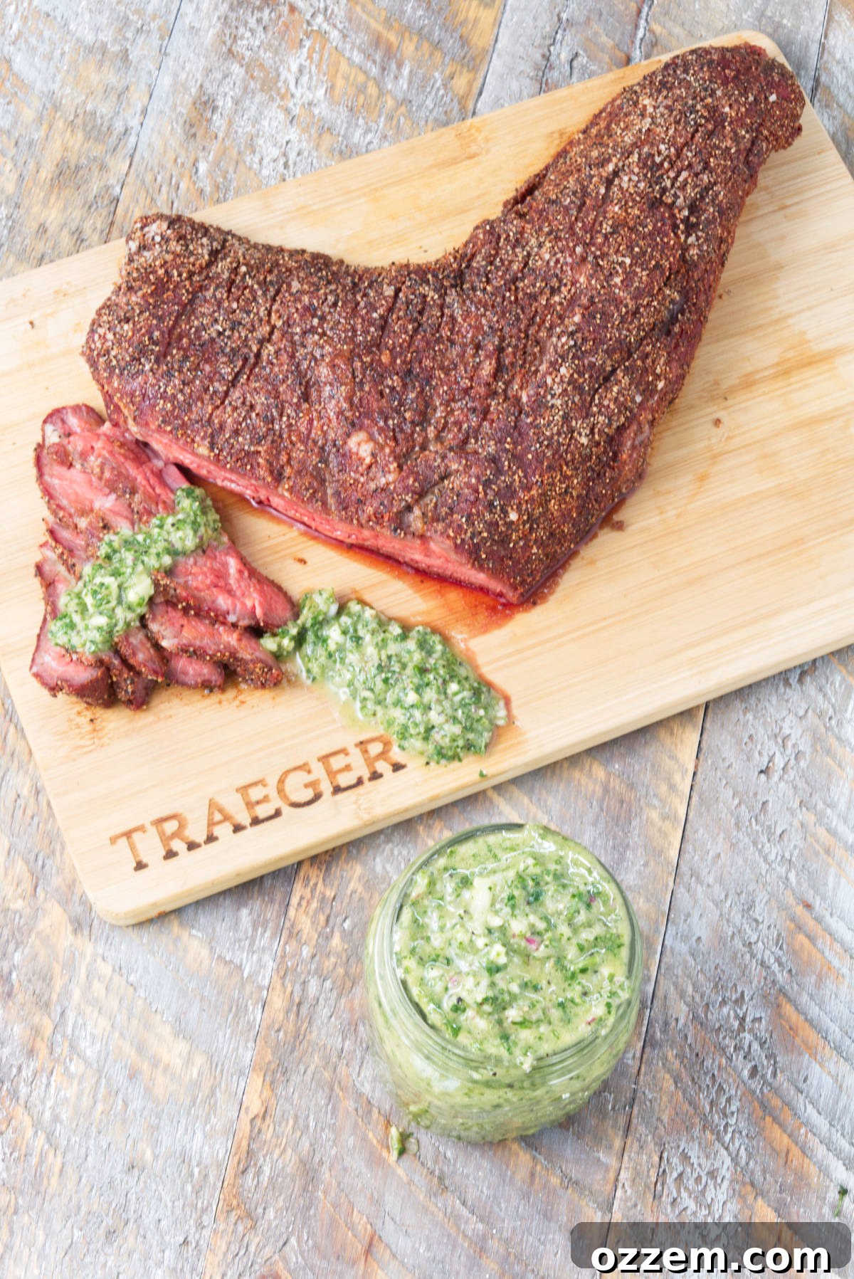 Sliced smoked tri-tip artfully arranged on a cutting board, generously drizzled with vibrant cilantro chimichurri, with a full mason jar of the sauce ready for serving.
