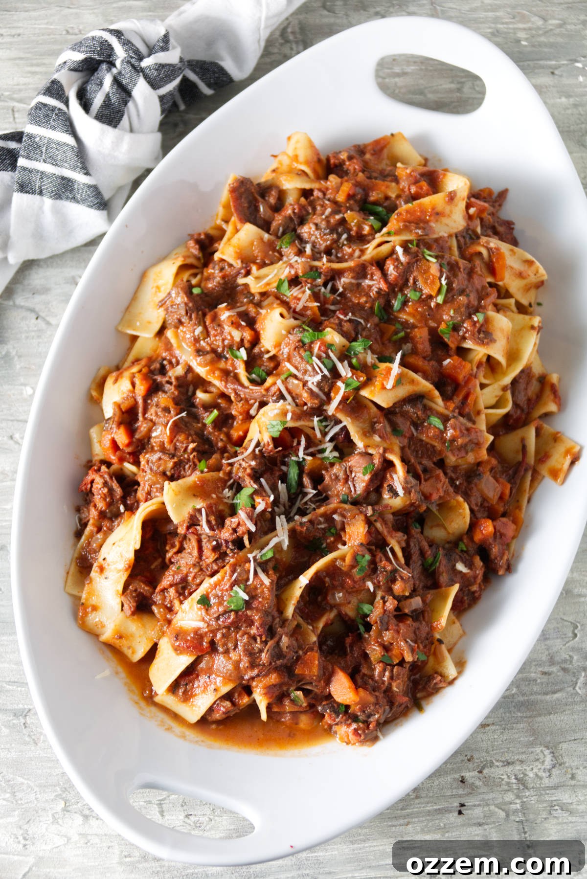 short rib ragu sauced served over pappardelle pasta in white bowl and finished with grated parmesan and parsley.