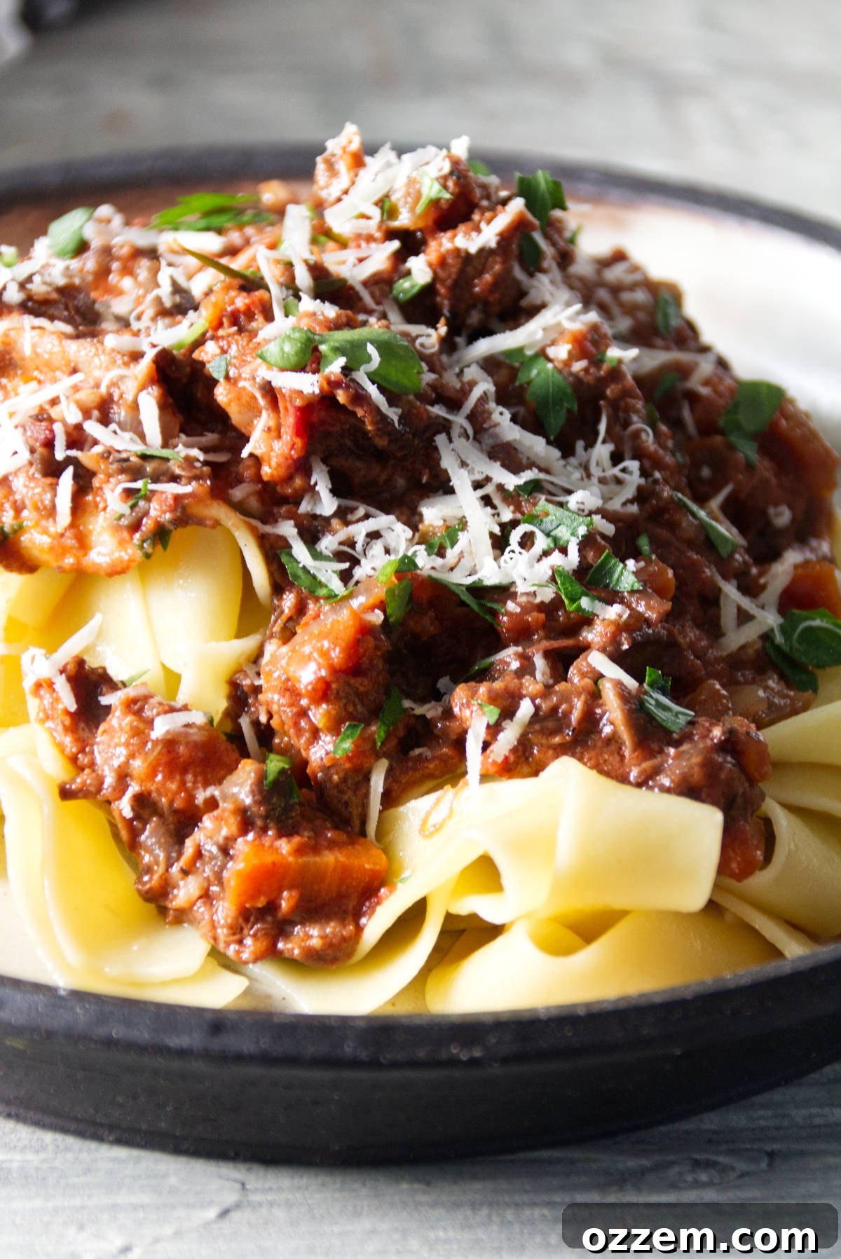 short rib ragu sauce served over pappardelle pasta on a plate and finished with grated parmesan and parsley.
