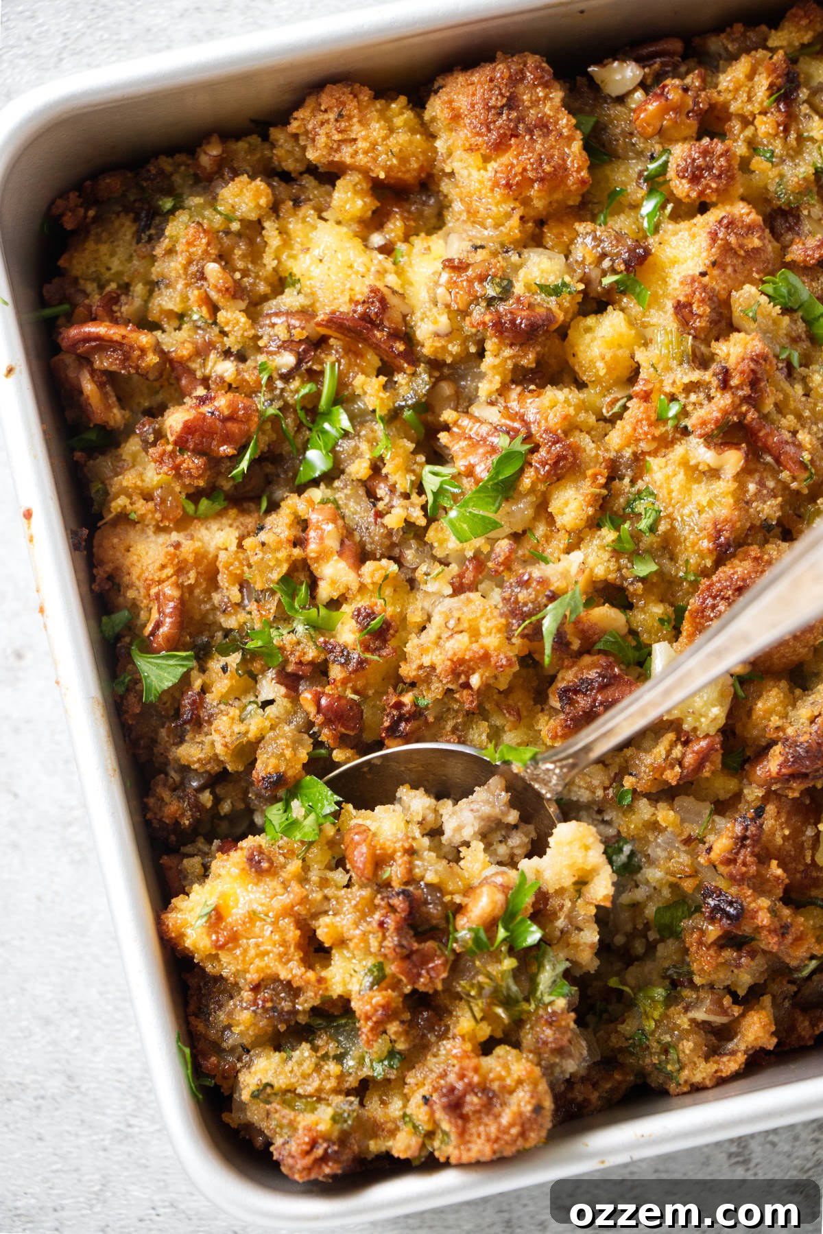 A serving spoon scoops rich, golden-brown air fryer stuffing from a festive baking dish, highlighting its crispy top and tender interior, flecked with sausage, herbs, and vegetables.