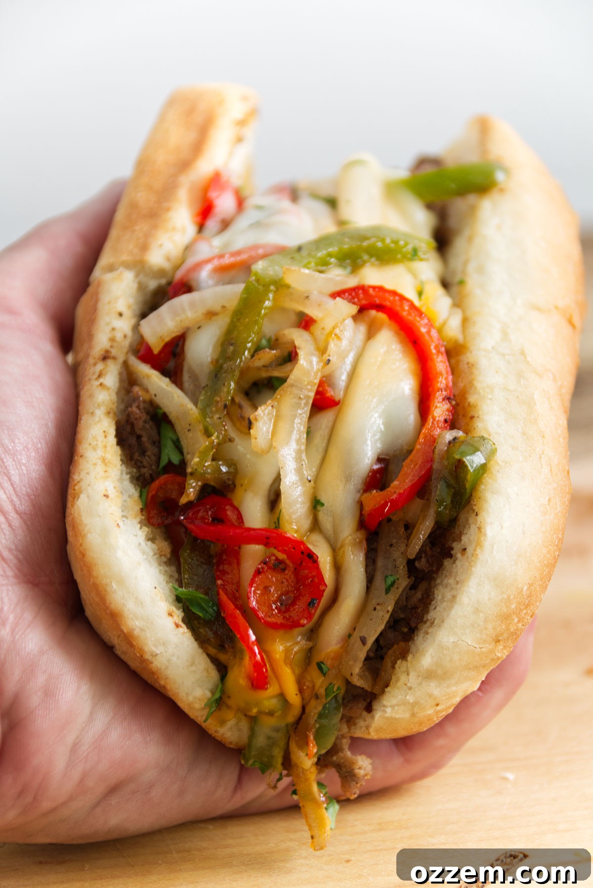 Sizzling Griddle Philly Cheesesteak 12 A hand holding a perfectly assembled Philly cheesesteak sandwich, showcasing its generous filling of onions, peppers, and an abundance of gooey Cheese Whiz and provolone cheese, ready for a satisfying bite.