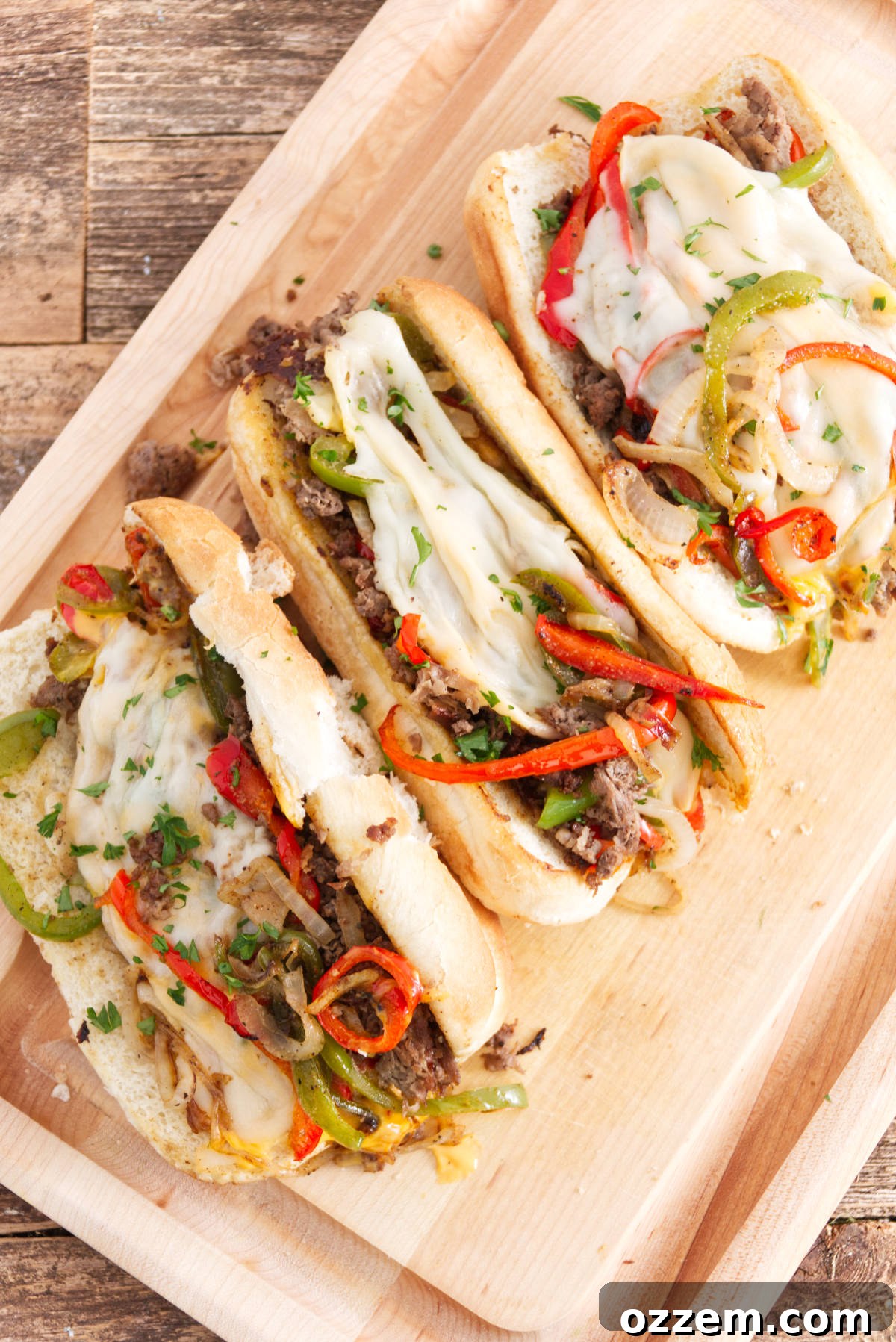 Sizzling Griddle Philly Cheesesteak 3 Three mouth-watering Philly cheesesteak sandwiches artfully arranged on a cutting board, each brimming with finely sliced steak, colorful red and green bell peppers, generous portions of melted provolone, and a decadent drizzle of Cheese Whiz.