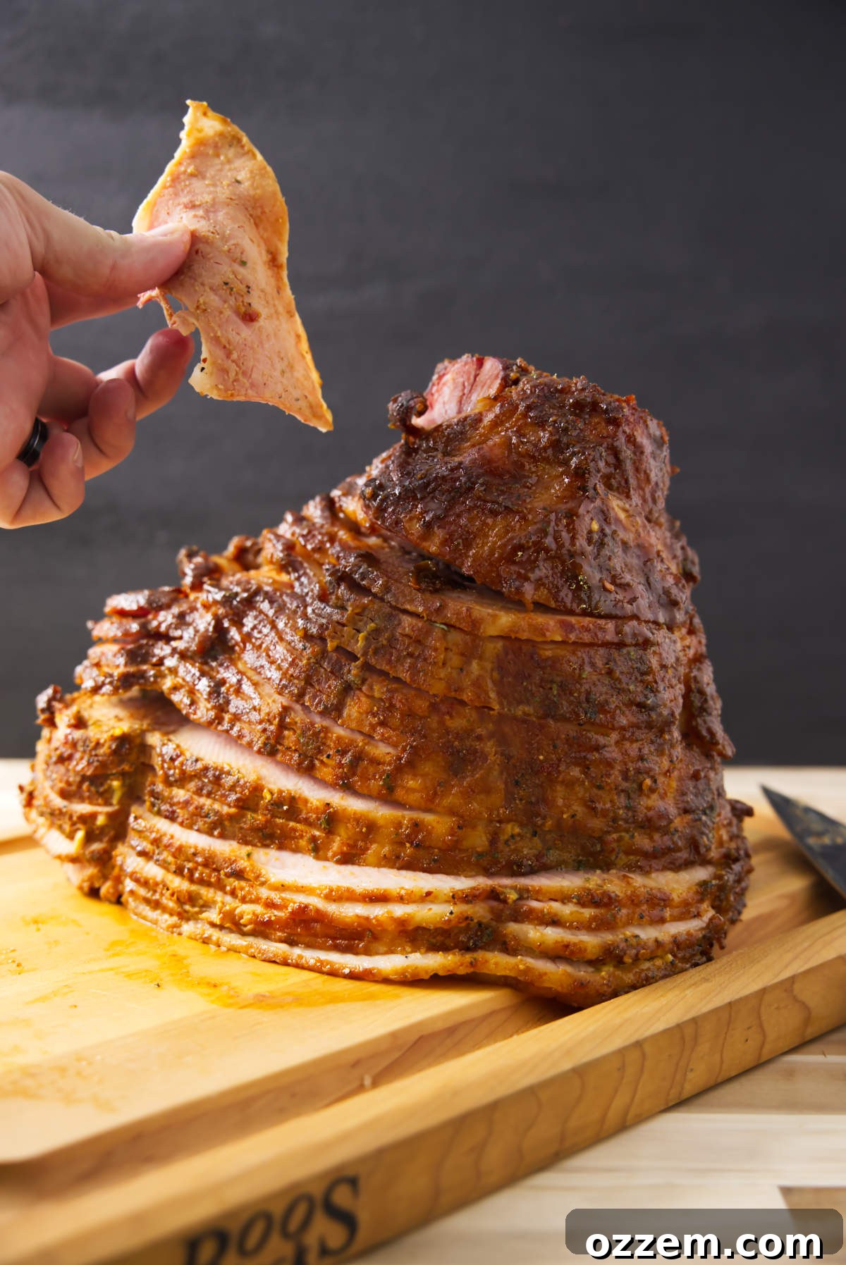 A hand holding a perfectly sliced piece of double smoked ham, with the remaining spiral ham glistening on a butcher block.