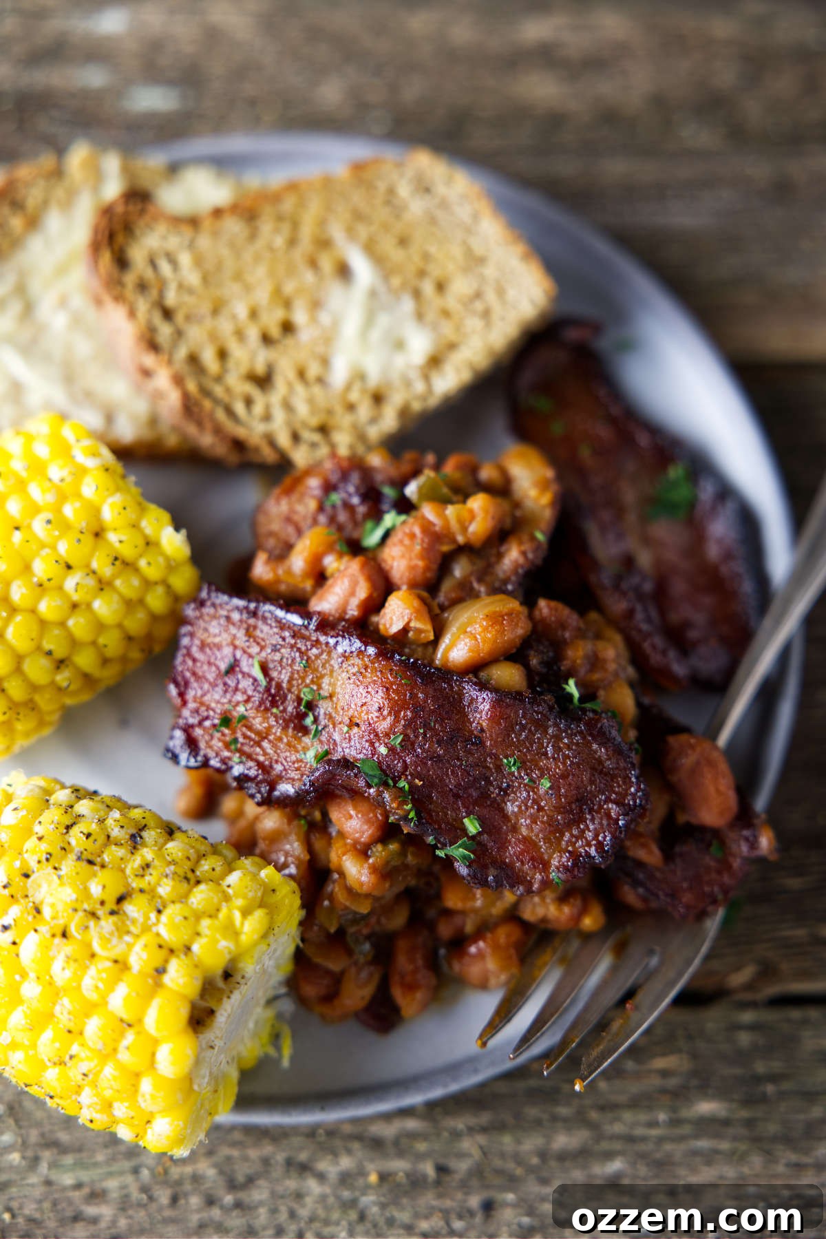 Pitmaster's Baked Beans 5 A plate featuring a generous serving of smoked baked beans, alongside grilled corn on the cob and a slice of crusty bread, ready for a summer meal.