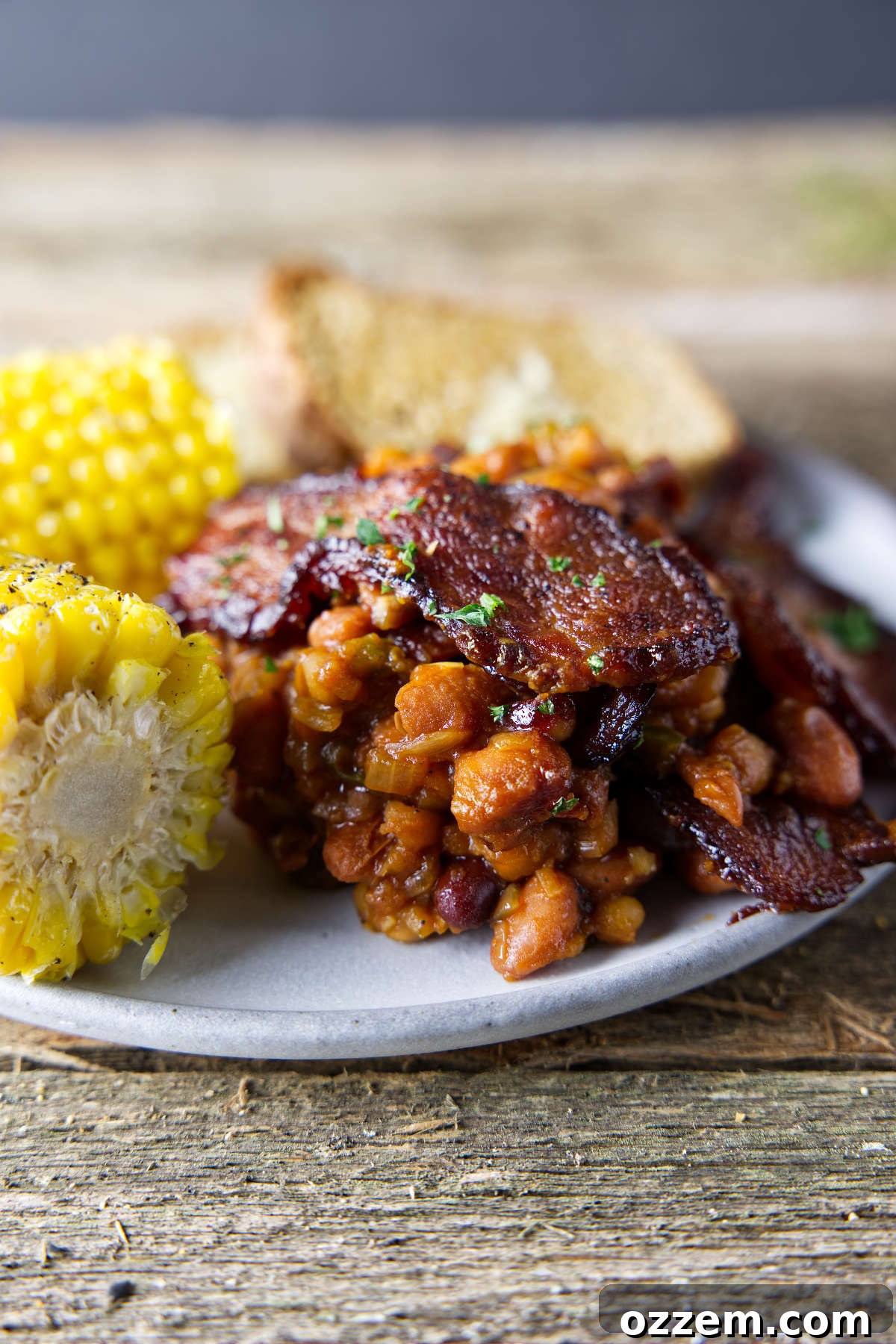 Pitmaster's Baked Beans 6 A plate featuring a generous serving of smoked baked beans, topped with a thick slice of crispy bacon, highlighting its rich texture and appetizing appeal.