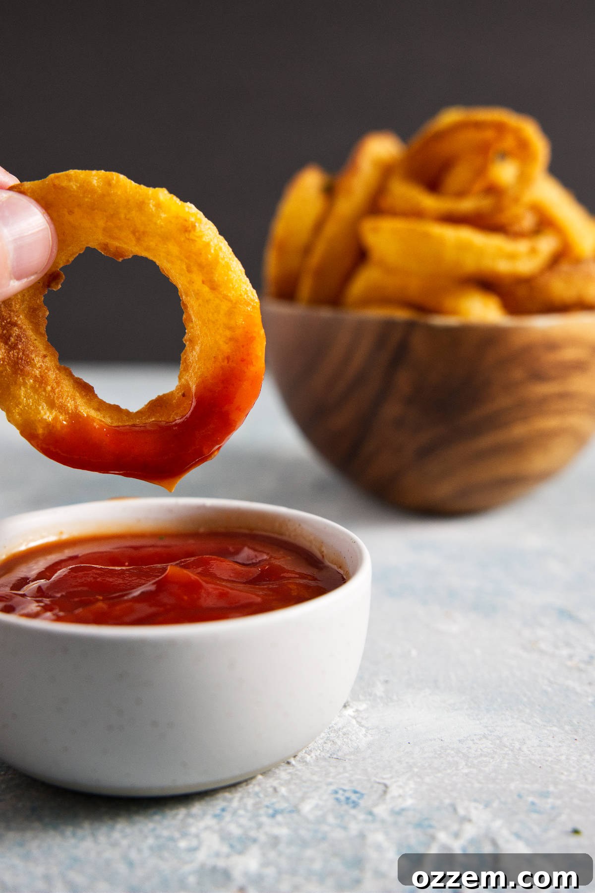 Crispy Air Fryer Frozen Onion Rings 6 Onion ring being dipped into ketchup with wooden bowl of onion rings in background