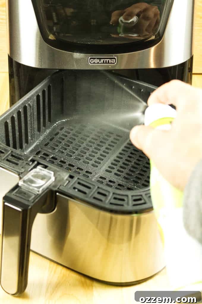 Air Fryer Mastery: Essential Tips and Tricks 4 Close-up shot of aerosol avocado oil being sprayed directly into the basket of a stainless steel and black air fryer, resting on a rustic wooden table.