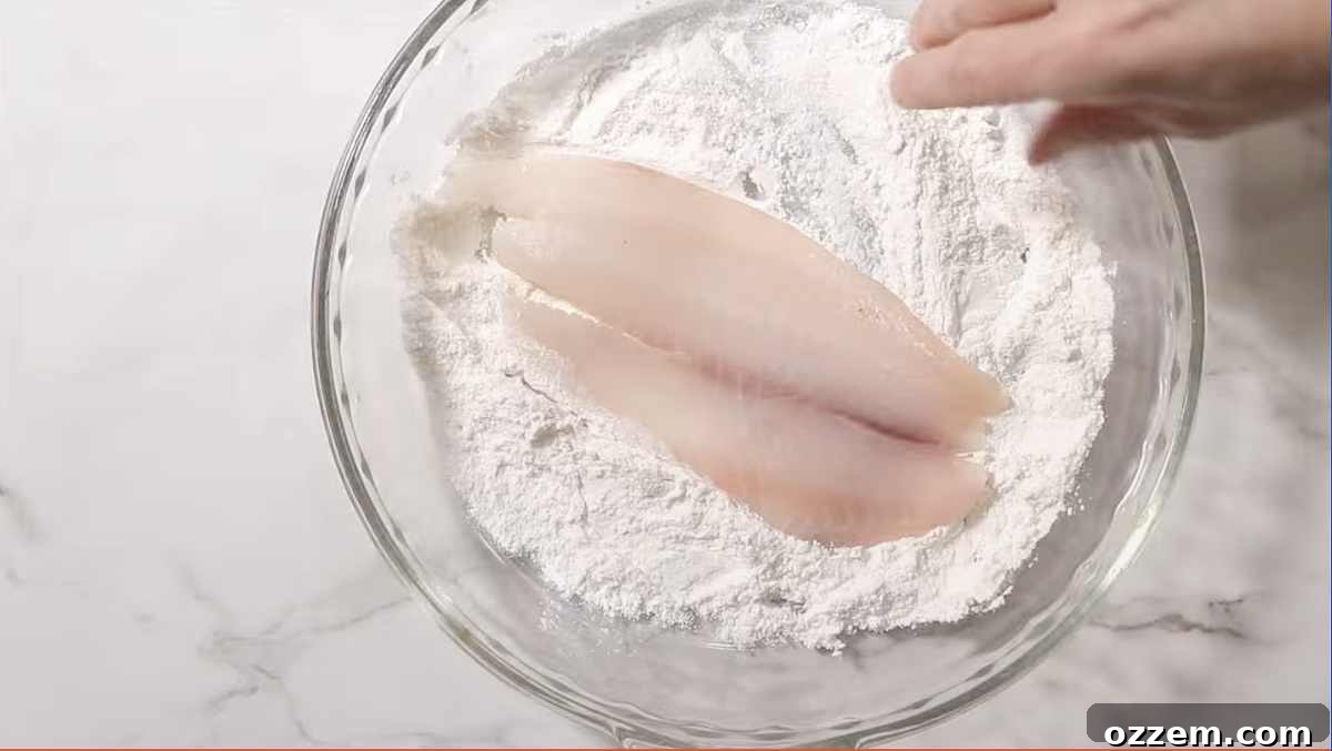 A hand dredging a tilapia fillet in a shallow dish of white flour, coating it evenly.