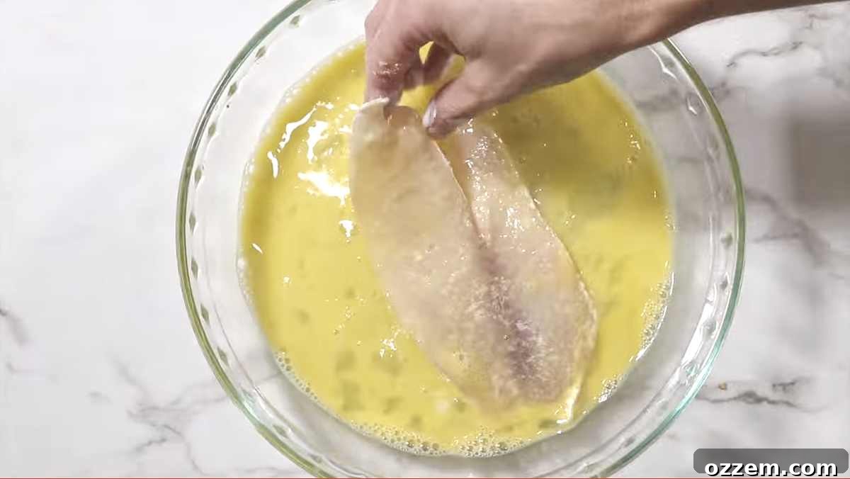 A tilapia fillet being dipped into a bowl of whisked egg wash, ensuring full coverage.