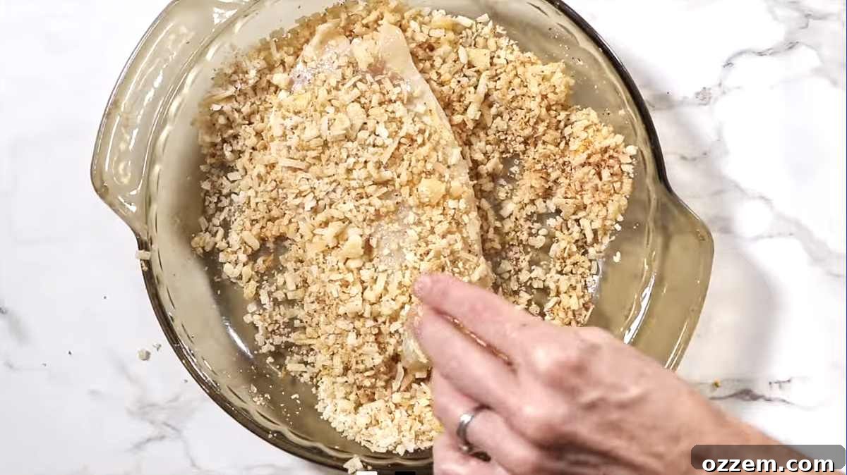 A hand pressing a tilapia fillet into a mixture of grated Parmesan cheese and spices to form a crispy coating.