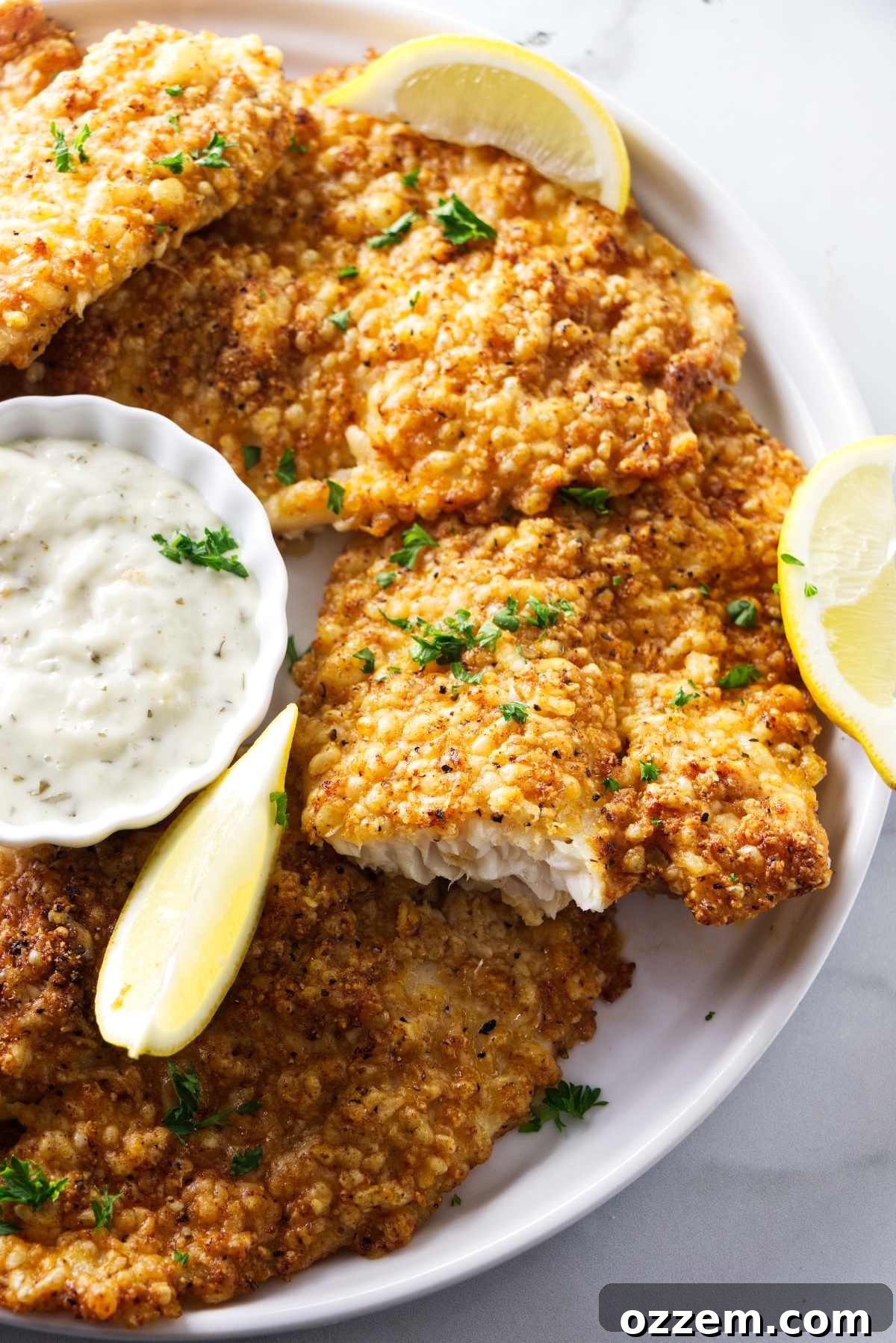 Several golden-brown, crispy air-fried tilapia fillets arranged on a platter, accompanied by lemon wedges and a small bowl of creamy tartar sauce.