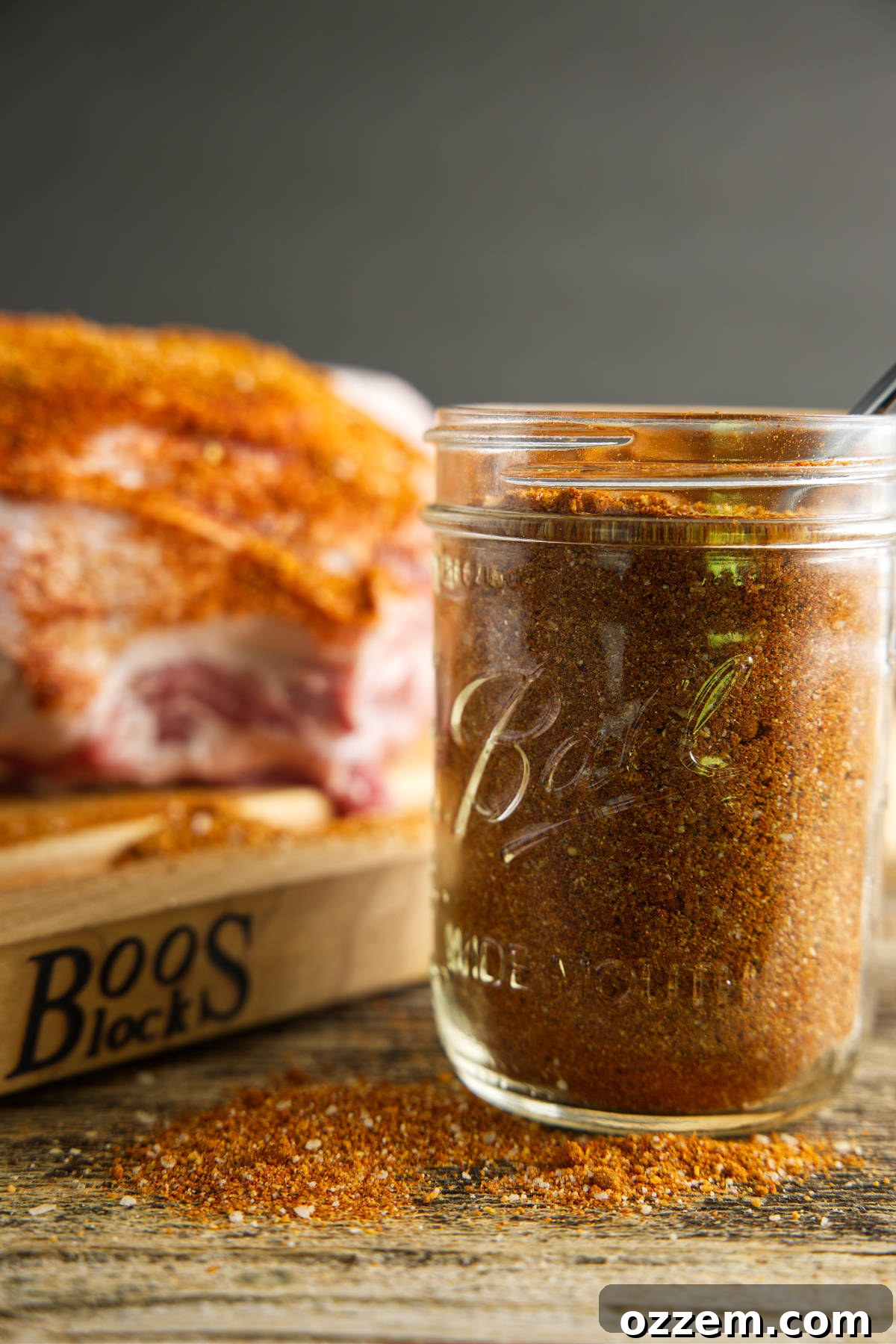 A glass jar filled with homemade pork butt rub, with a perfectly seasoned pork butt visible in the soft-focus background, ready for smoking.