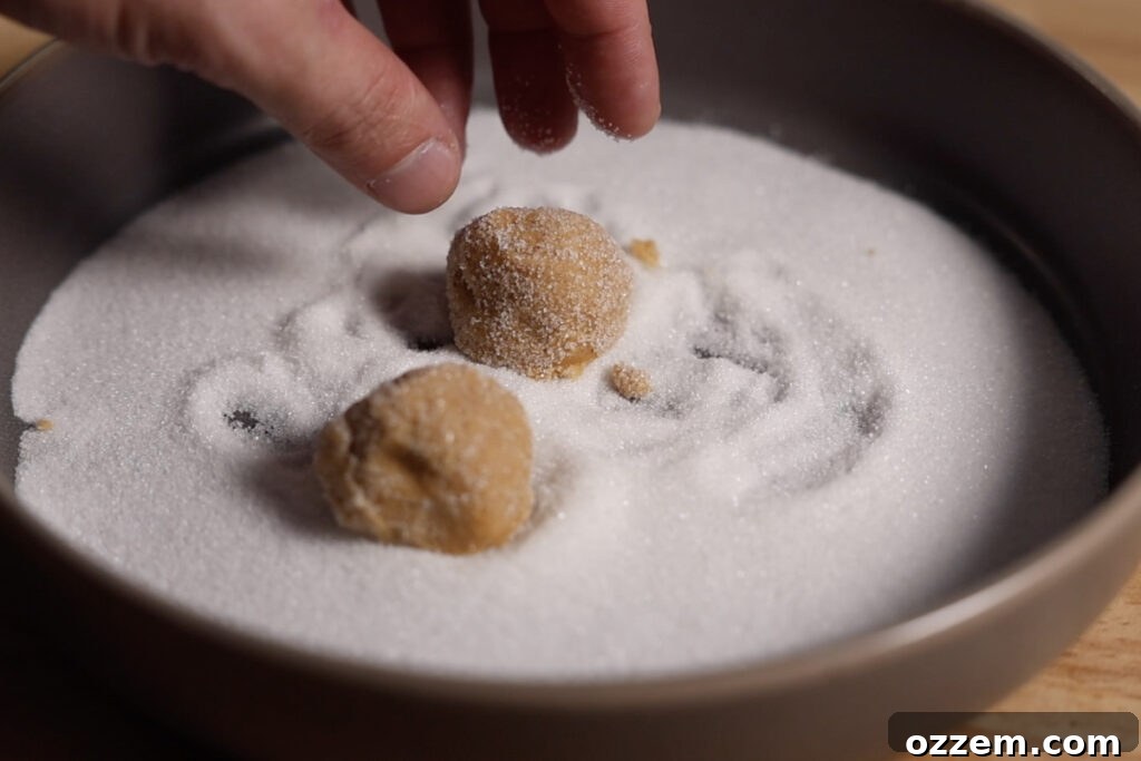 Uniform cookie dough balls being meticulously rolled in granulated sugar, preparing them for baking with a sweet, glistening coating.