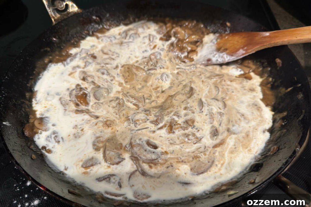Heavy cream being slowly poured into the mushroom and onion mixture in a frying pan, to form a creamy sauce.