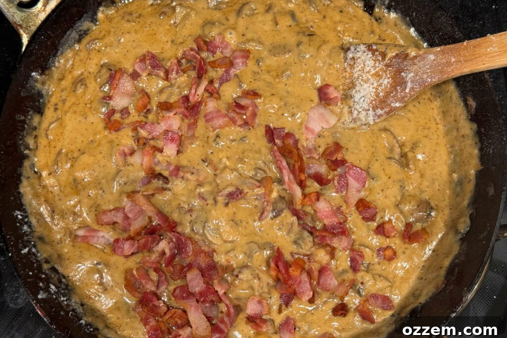 Crispy bacon and blanched green beans being folded into the creamy sauce within a large fry pan.