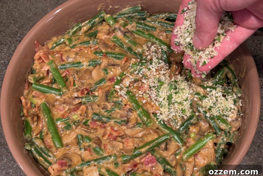 A green bean casserole dish being topped generously with a golden breadcrumb mixture before smoking.