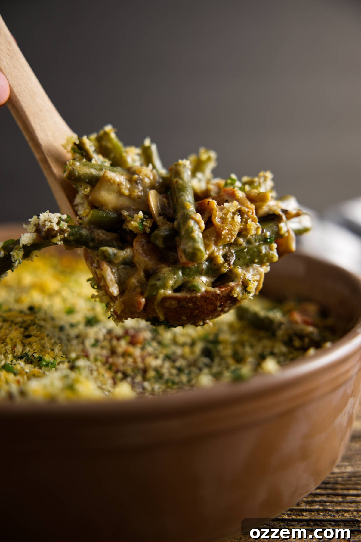 A large scoop being taken from a casserole dish of freshly smoked green bean casserole, revealing the creamy interior, tender green beans, bacon, and mushrooms, with a crispy breadcrumb topping.
