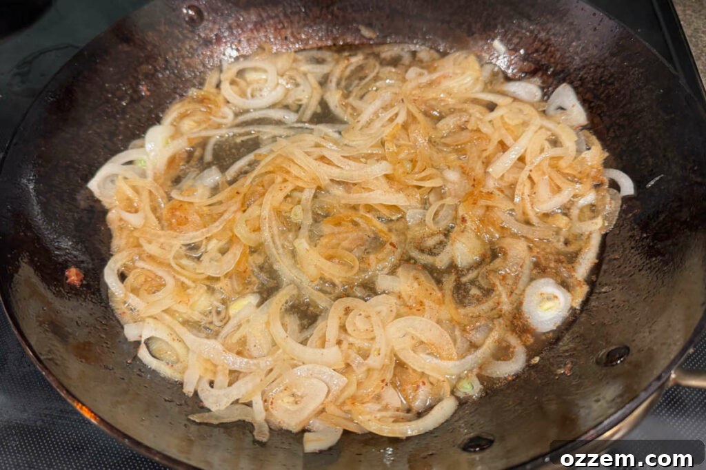 Sliced white onions sautéing in a dark fry pan on a stovetop, becoming translucent and soft.