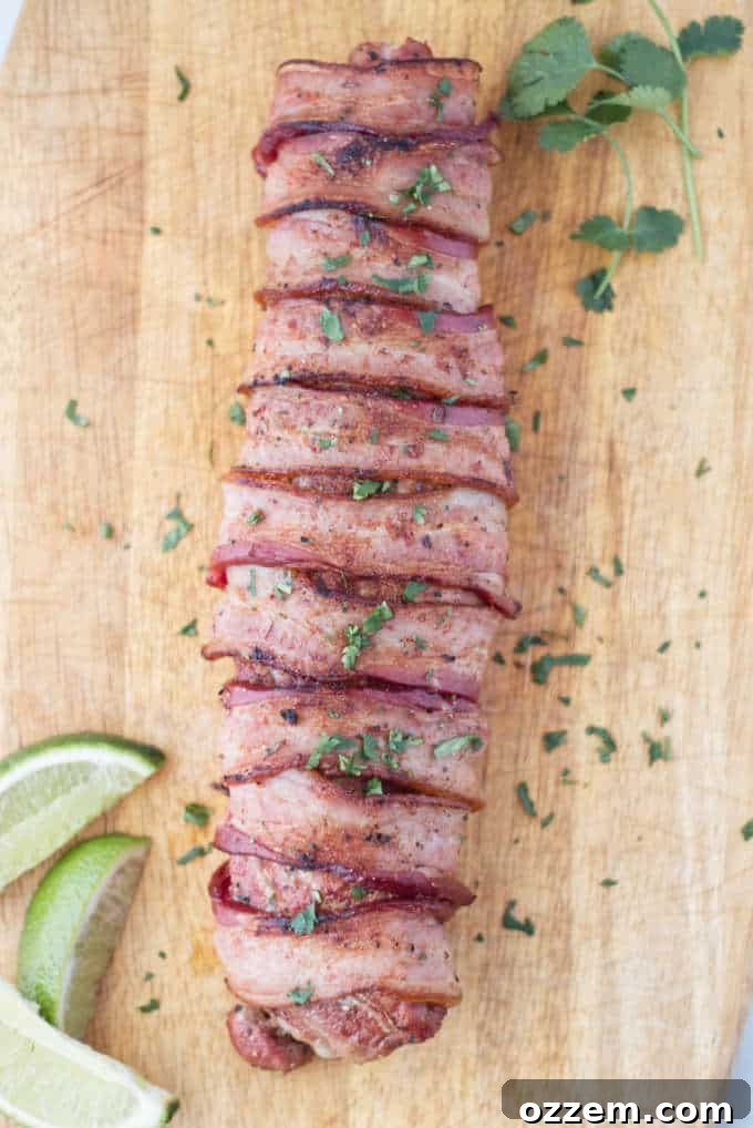 Traeger Smoked Bacon-Wrapped Pork Tenderloin 2 12 slices of bacon wrapped around a pork tenderloin, laying on a wood cutting board with 3 slices of lime and some sprigs of cilantro and chopped cilantro sprinkled on top.