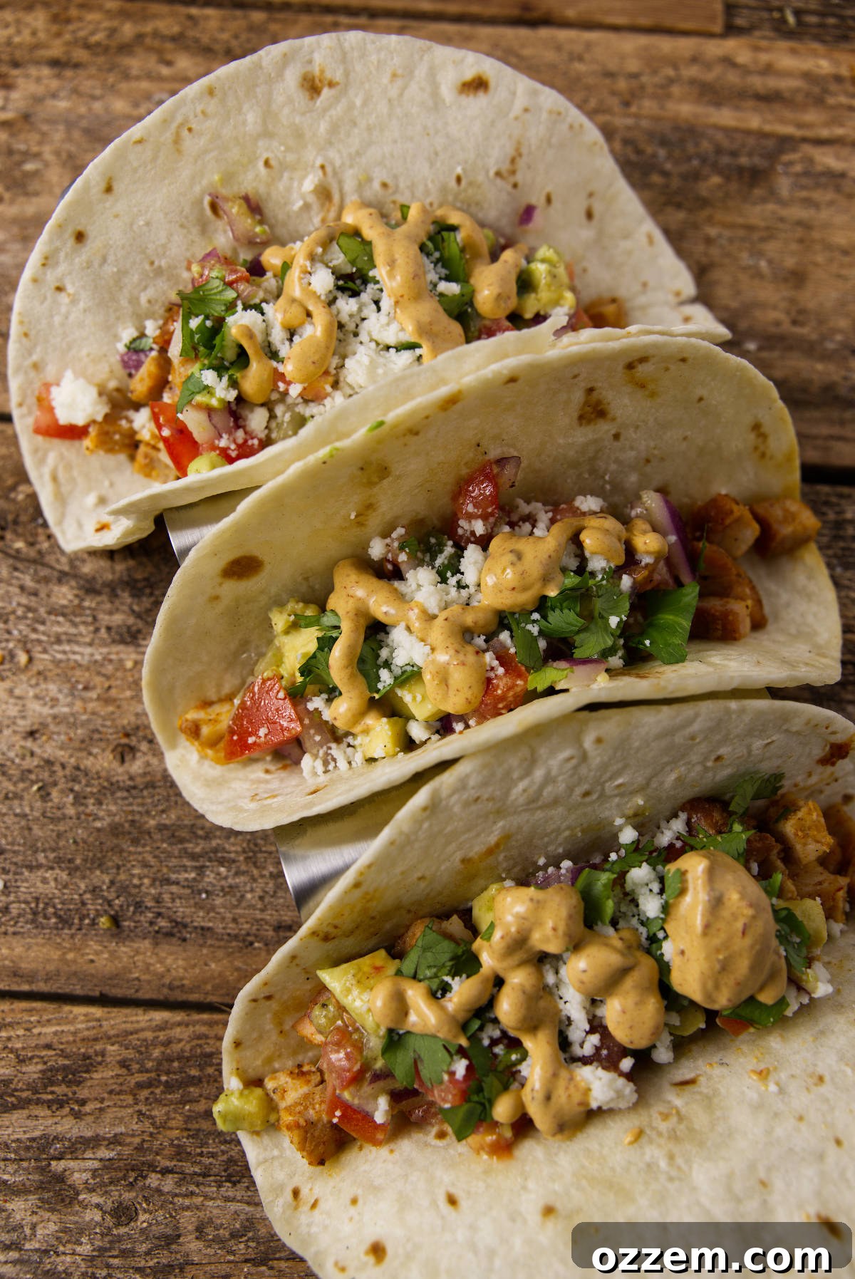 Pork Loin Taco Remix 2 Overhead view of three gourmet pork loin tacos, generously topped with vibrant avocado salsa, creamy chipotle lime crema, and crumbled queso fresco, presented on a rustic wooden board.
