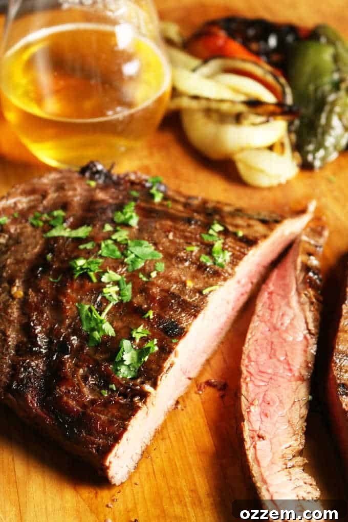 Sliced grilled flank steak on a wooden board with a glass of beer and fresh vegetables in the background