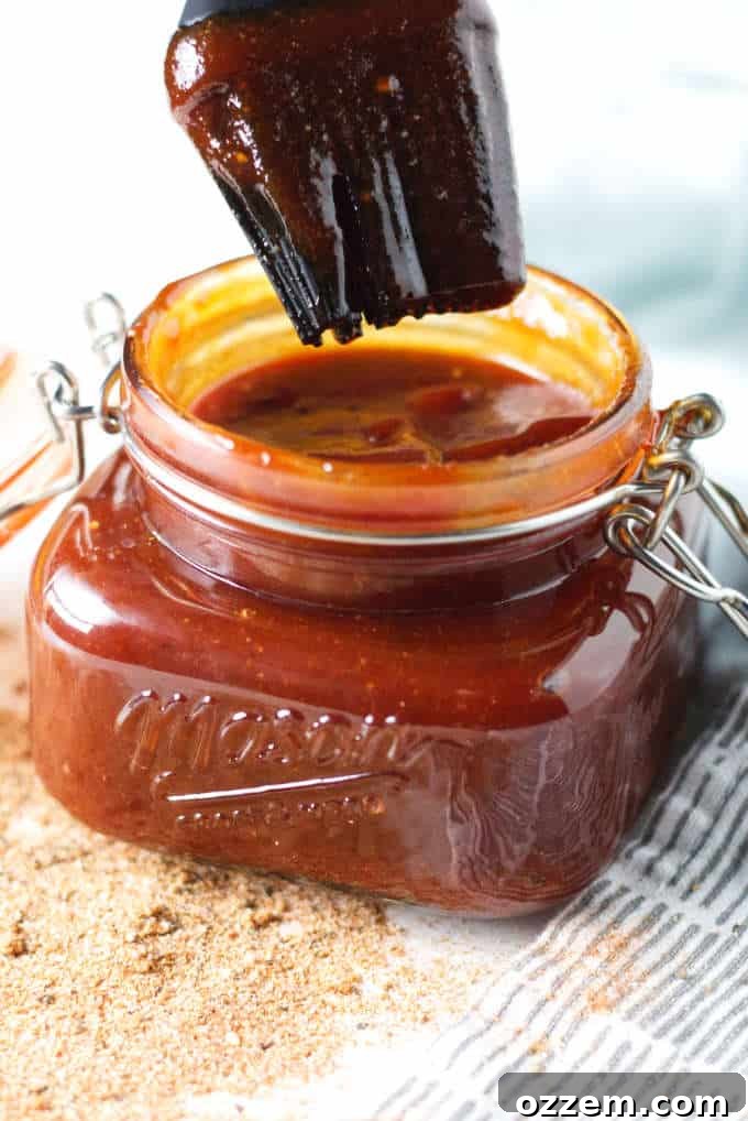 A mason jar filled with rich, dark Kansas City-style BBQ sauce, placed on a grey and white checkered napkin. Loose spices are scattered on the table in front, hinting at the sauce's flavorful ingredients.