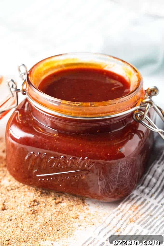A mason jar filled with freshly made Kansas City-style BBQ sauce, gleaming on a grey and white napkin with a sprinkle of seasoning. The background hints at a bustling kitchen, ready for grilling.