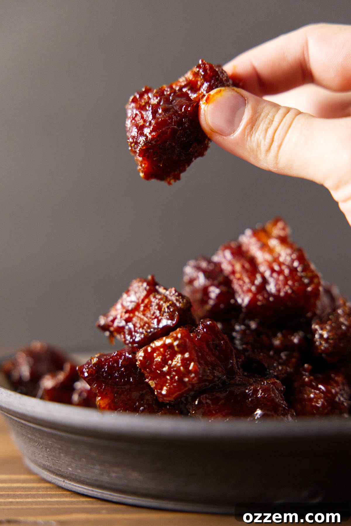 A hand holding up a glistening, caramelized piece of smoked pork belly burnt end, showcasing its tender texture and delicious coating.