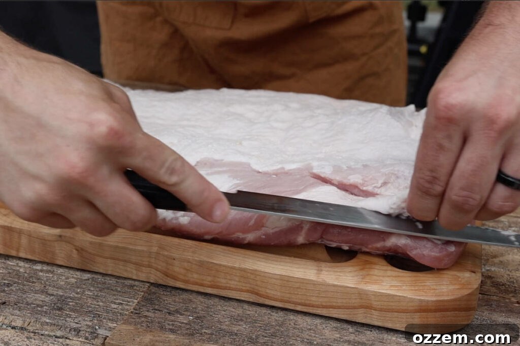A close-up of a chef's hands carefully trimming excess fat from a large slab of pork belly on a wooden cutting board.