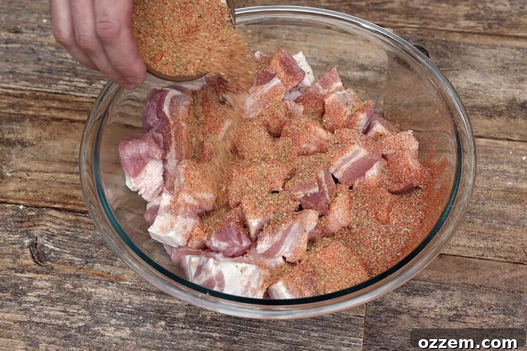A generous amount of spice rub being poured over cubed pork belly in a mixing bowl, ensuring thorough coating.