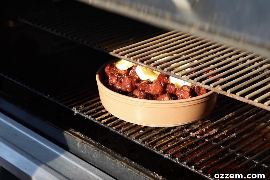 A pan of sauced pork belly burnt ends smoking on a Traeger grill, covered with foil, for the final tenderizing phase.