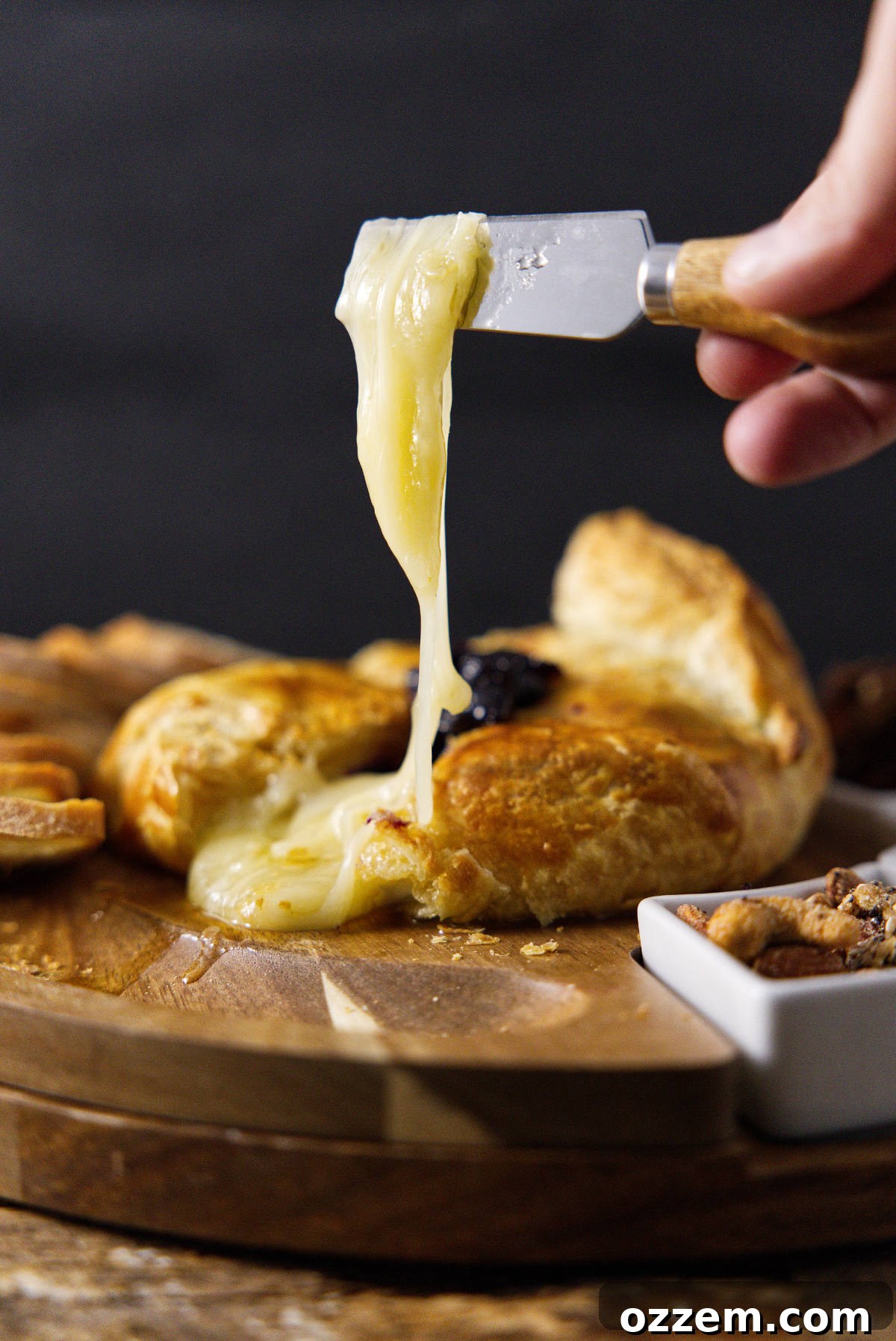 A cheese knife carefully lifting a piece of perfectly melted brie cheese, oozing enticingly from its crispy puff pastry shell.