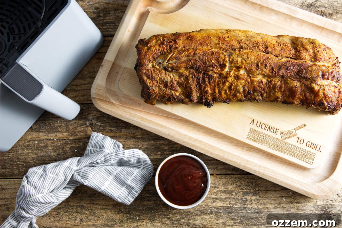 Slab of pork ribs resting on a wooden cutting board, with an air fryer basket and a bowl of rich BBQ sauce nearby.