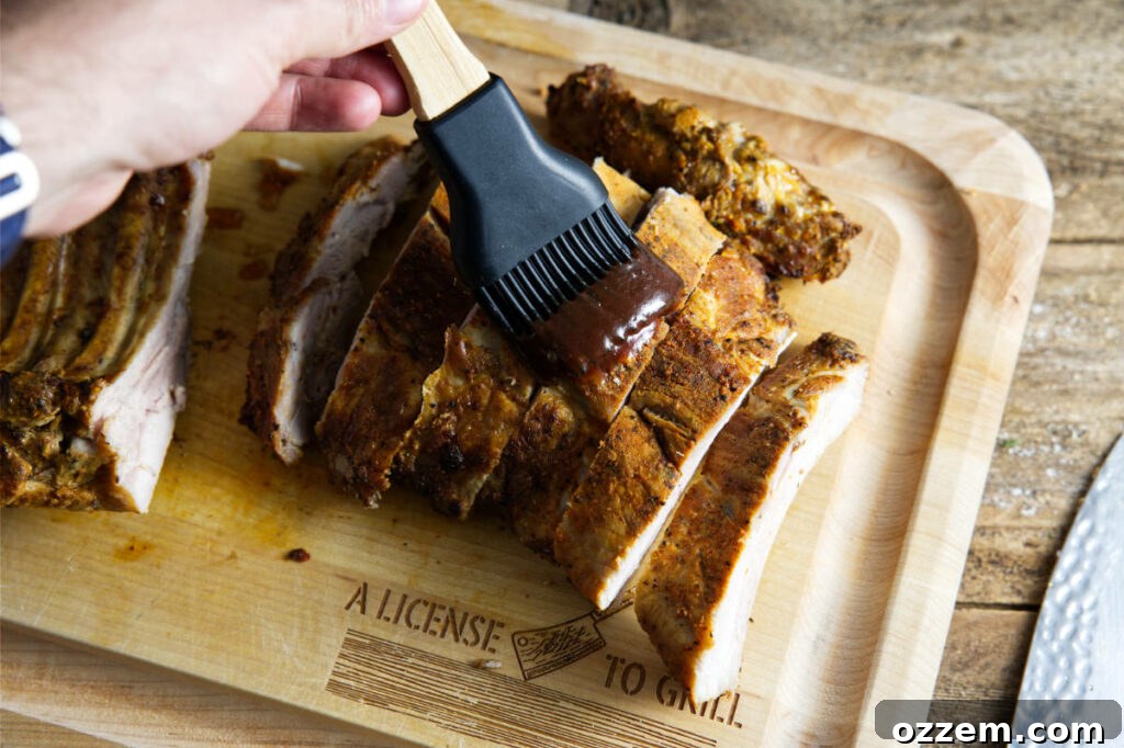 A hand brushing rich BBQ sauce onto pork ribs arranged on a cutting board.