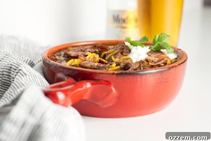 small red pot filled with brisket chili with cheese and sour cream and a garnish of cilantro, modelo beer bottle with glass of beer in background and napkins in foreground