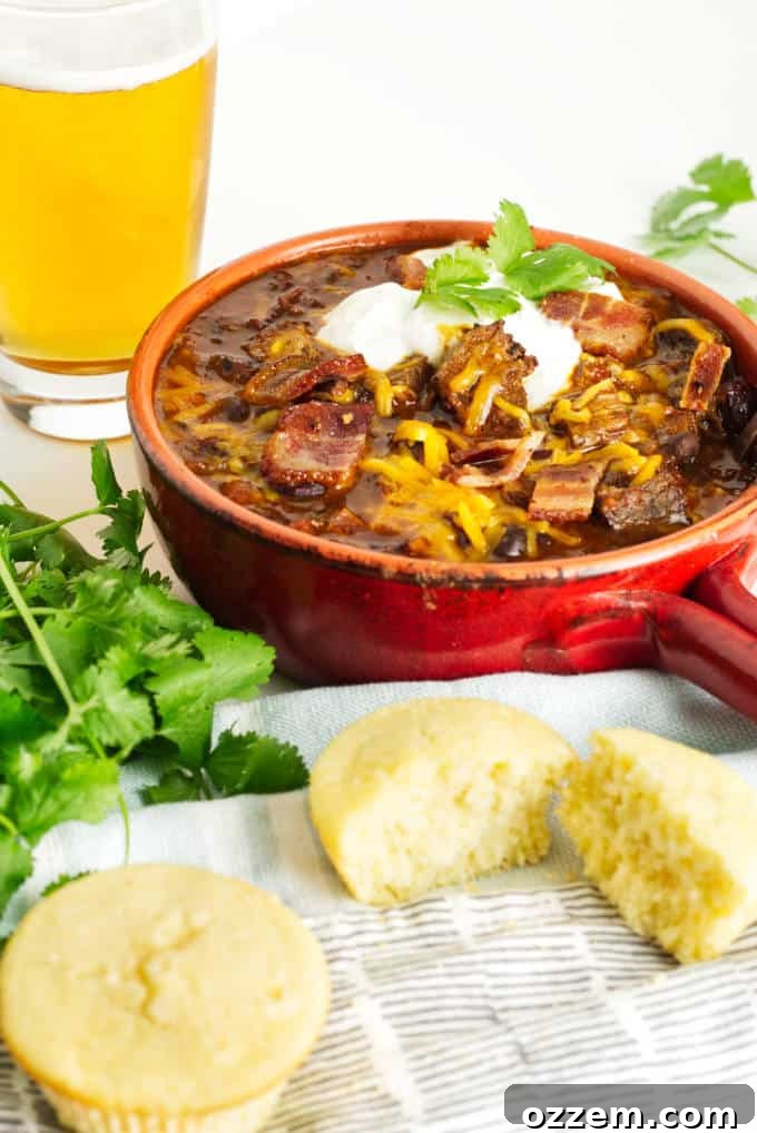 small red pot filled with brisket chili with cheese and sour cream and a garnish of cilantro, glass of beer in the background and cornbread in the foreground