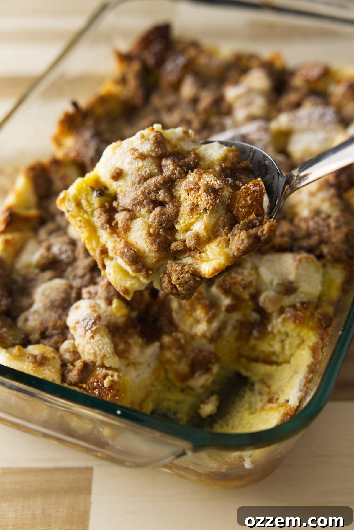 A large metal spoon scooping a portion of golden-brown baked French toast casserole from a baking dish, highlighting the soft interior.