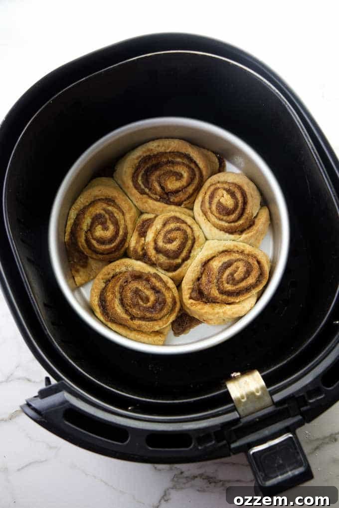 Hot cinnamon rolls nestled in a cake pan inside an air fryer basket, with delicate steam gently rising, indicating their fresh-baked warmth.