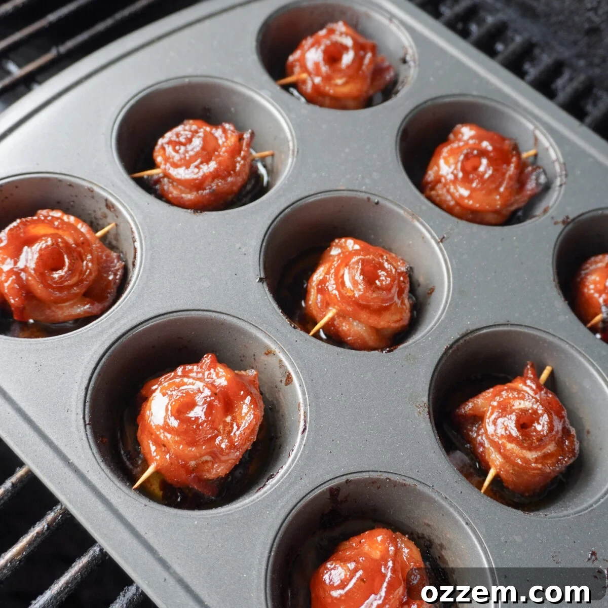 Grilled bacon roses cooking in a muffin pan on an outdoor grill, with smoke rising.