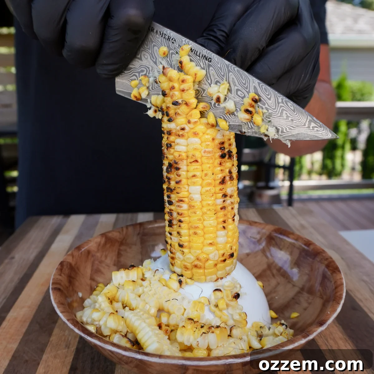 Roasted corn on the cob upright with kernels being sliced off by knife into bowl.