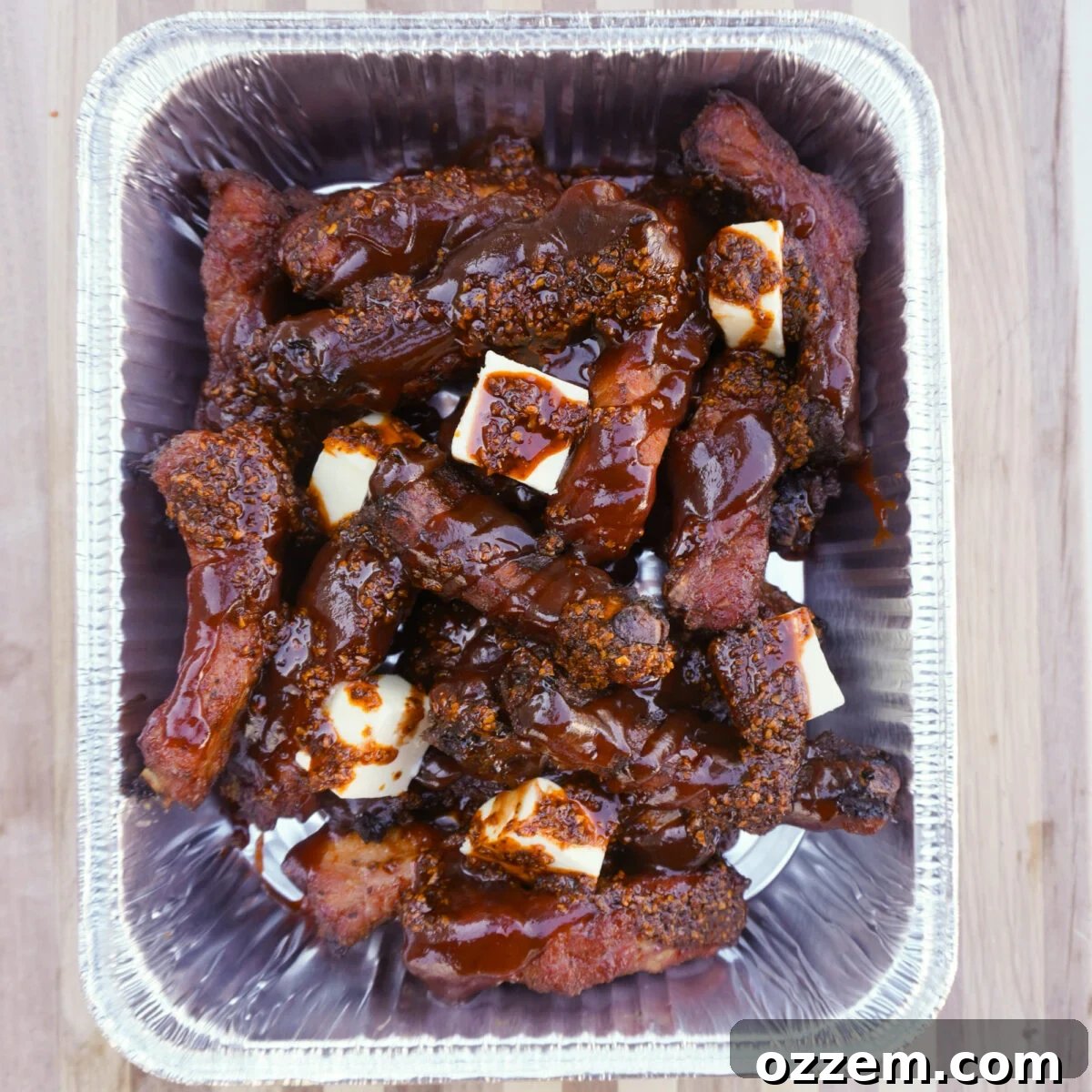 Ultimate Chili Crunch Ribs 8 Smoked ribs covered in a rich mixture of BBQ sauce, chili crunch, and butter in an aluminum foil pan.
