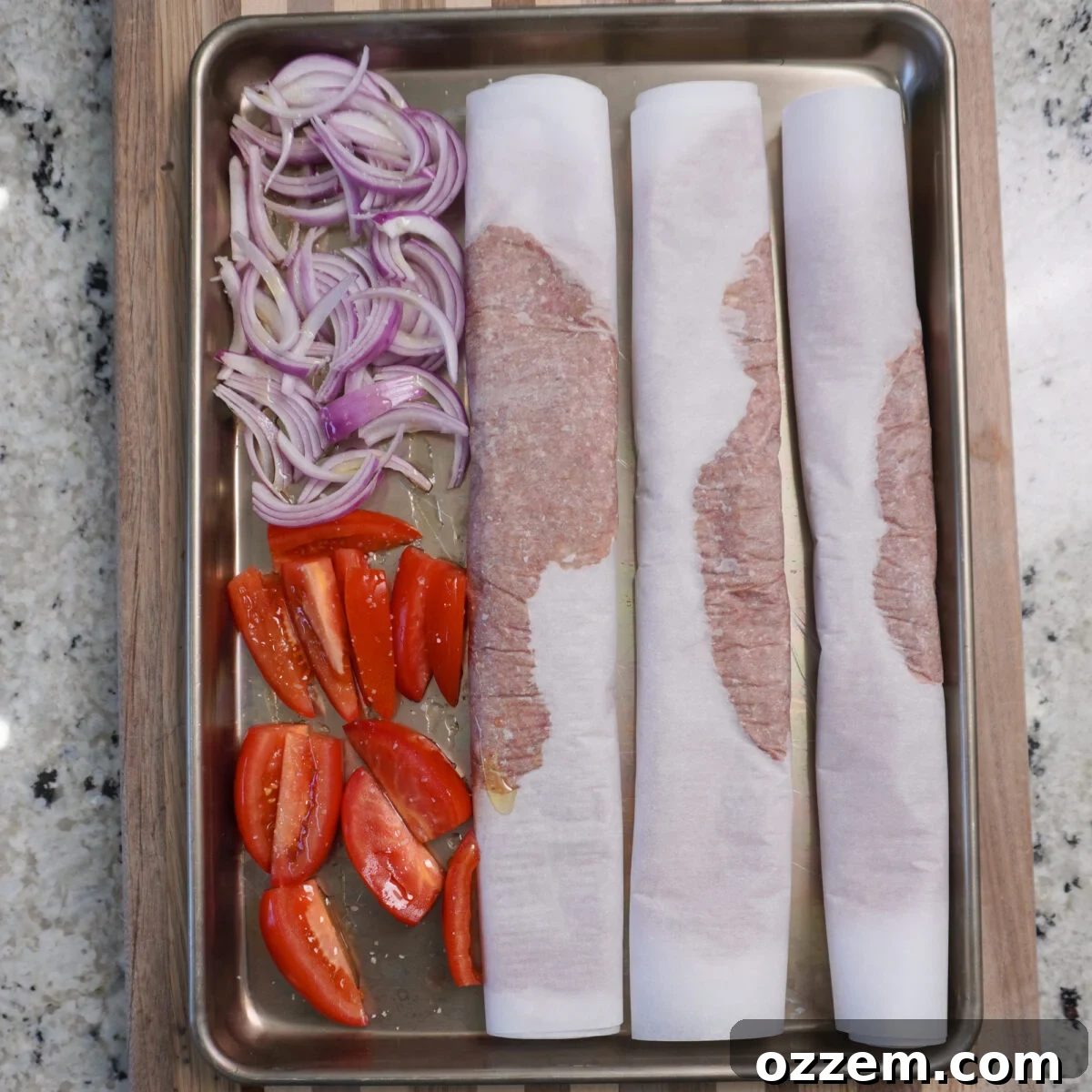 A sheet pan loaded with prepared beef kebabs, sliced Roma tomatoes, and red onions, lightly drizzled with olive oil, ready for baking.