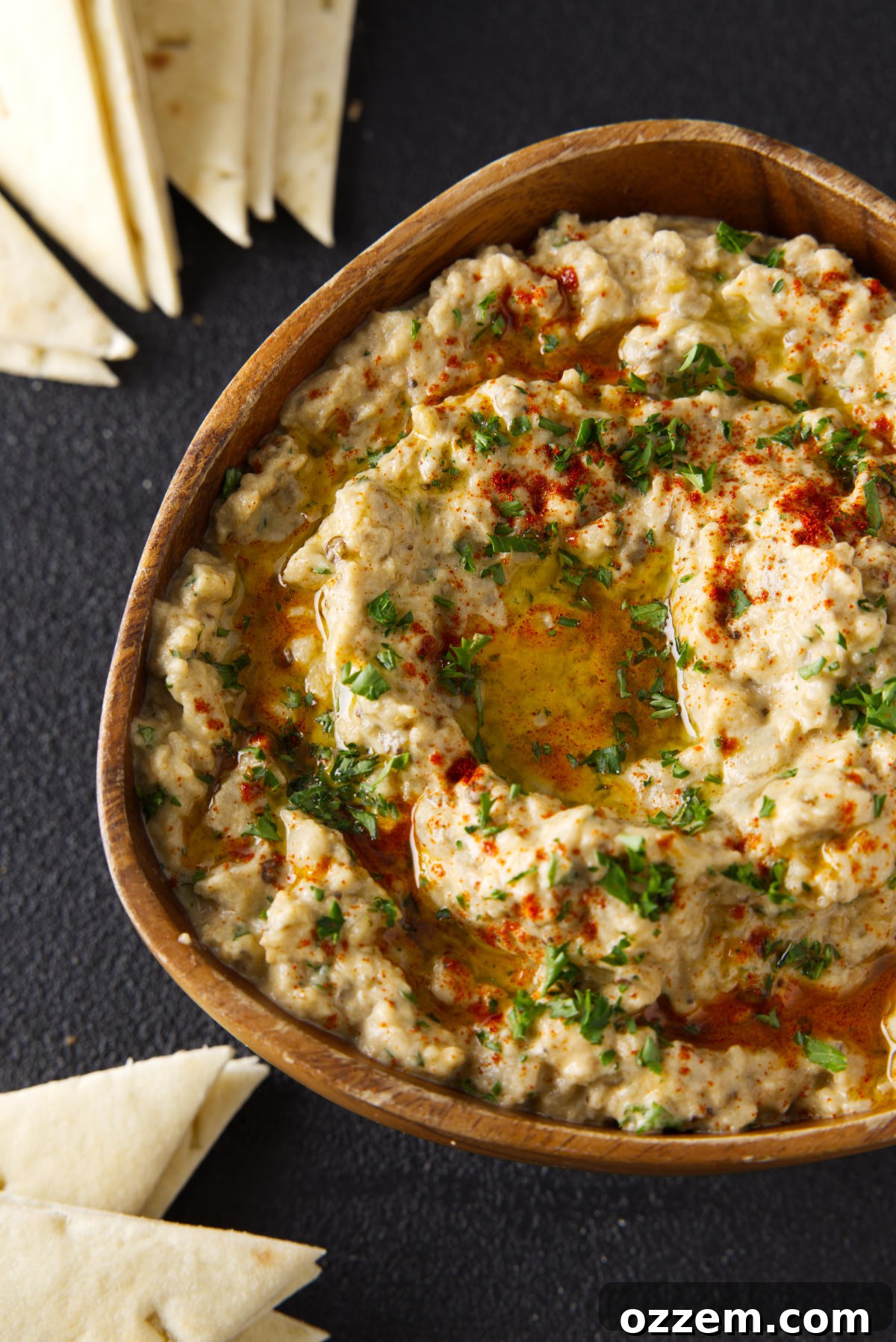 Overhead picture of smoked baba ganoush in a wooden bowl surrounded by slices of pita bread. The dip is garnished with fresh parsley and a drizzle of olive oil, showcasing its rich texture and appealing presentation.