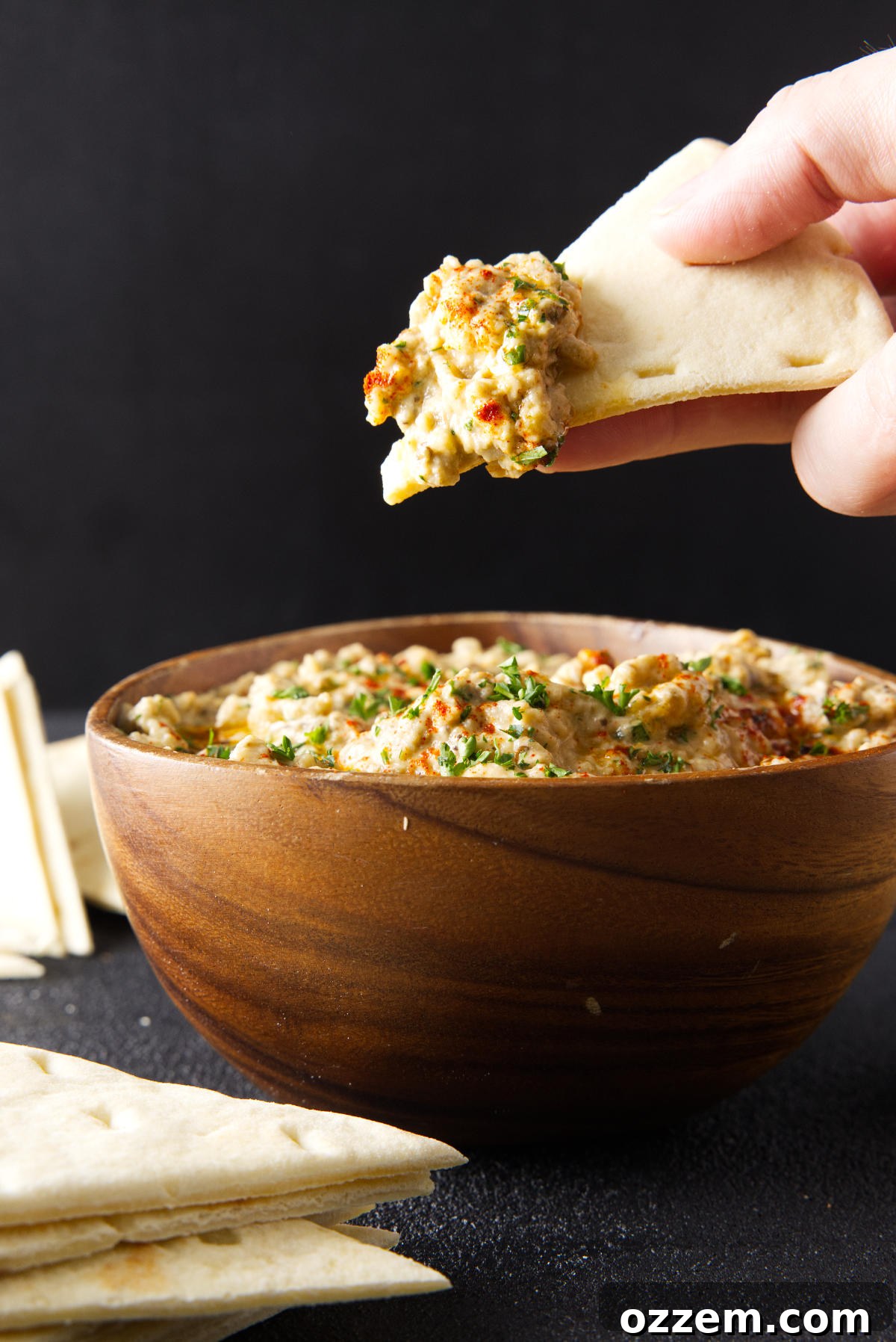 Dipping slice of pita bread in to a wooden bowl filled with baba ganoush, garnished with olive oil and parsley.